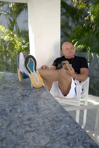 Shawn sits relaxed in a white chair with feet up on a stone table, holding a smartphone outdoors.