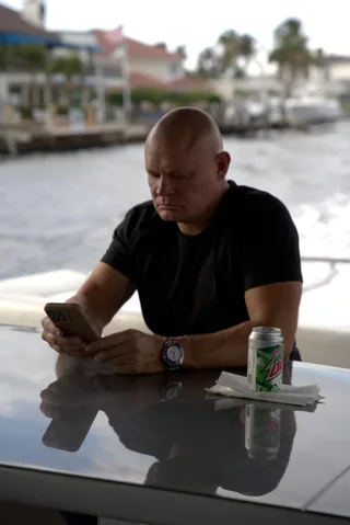 Shawn looks down at a smartphone while sitting at an outdoor table by the water.