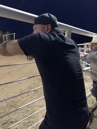 Shawn leans against a fence at a rodeo arena at night.
