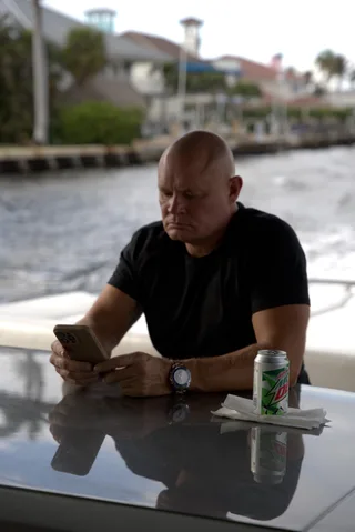 Shawn looks down at a phone while sitting at a table on a boat with a beverage can nearby.