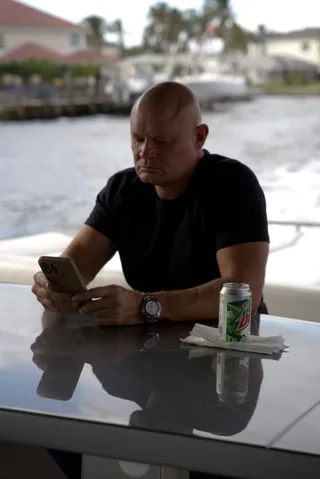 Shawn sits at a glossy table outdoors near water, looking down at a phone with a drink nearby.