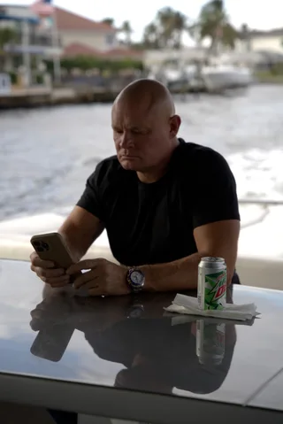 Shawn looks down at a phone while sitting at a table outdoors.