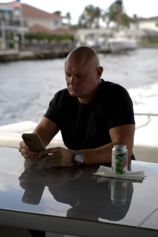 Shawn looks at a phone while sitting at a table outdoors near water.