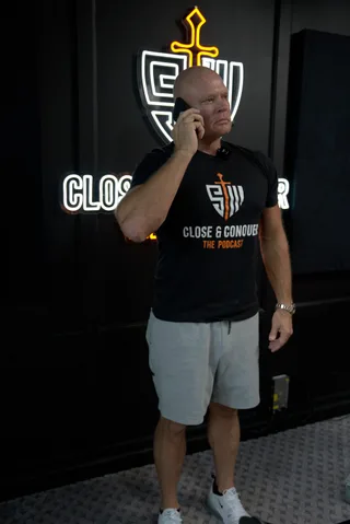 Shawn holds a phone to his ear while standing in a podcast studio with a neon logo in the background.
