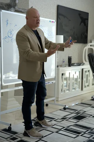 Shawn gestures with hands while teaching at a whiteboard in a modern room.