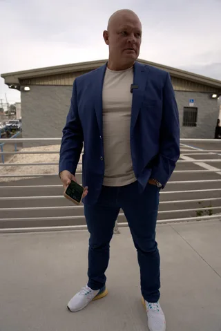 Shawn holds a phone while standing outdoors in a blue jacket and sneakers.