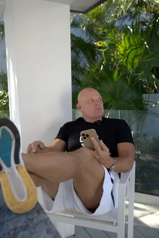 Shawn sits relaxed in a chair on a sunny patio holding a smartphone with feet up on a table.