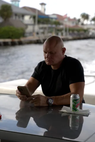 Shawn sits at a table looking down at a smartphone outside near the water.