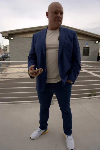 Shawn holds a phone while standing outdoors in a blue jacket and sneakers.