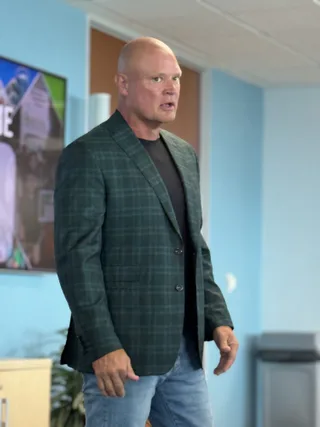Shawn stands in a plaid blazer and jeans, speaking in an office setting.