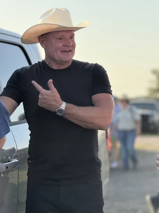 Shawn wears a cowboy hat while pointing outdoors beside a vehicle.