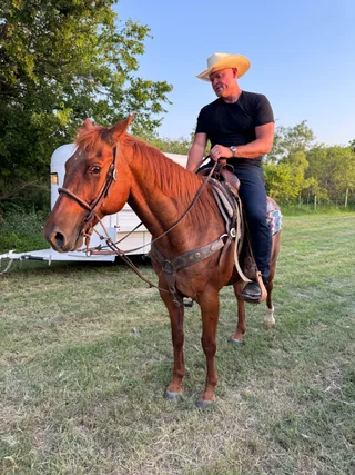 Shawn wears a cowboy hat while riding a chestnut horse in an open field near a horse trailer.