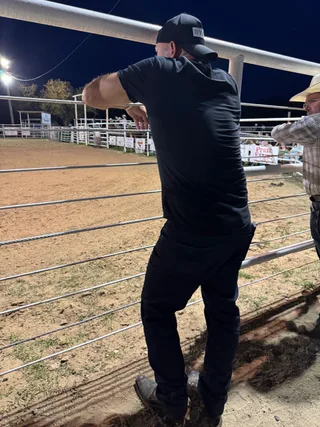 Shawn stands with his back to the camera, leaning on a metal fence at an outdoor rodeo arena at night.