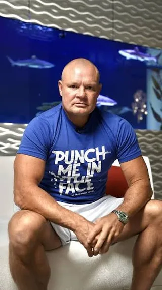 Shawn in a blue t-shirt sits on a white sofa in front of a shark aquarium.
