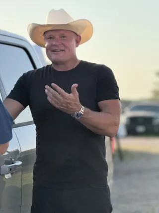 Shawn wearing a cowboy hat gestures while talking outside near a vehicle.