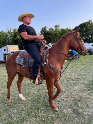 Shawn wears a cowboy hat while riding a horse in a grassy outdoor field.