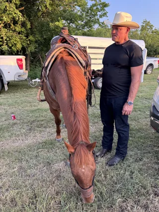 Shawn stands next to a saddled brown horse grazing in a grassy field.