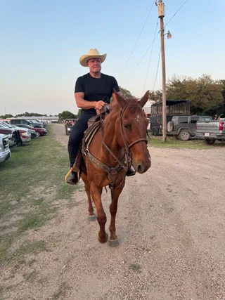 Shawn rides a brown horse along a dirt path outdoors.