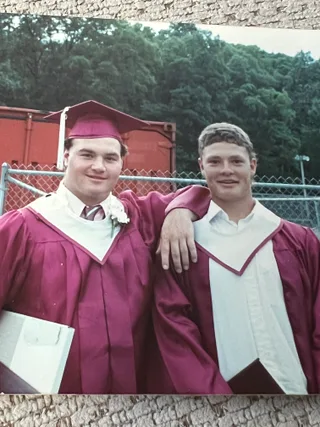 Shawn wearing a graduation gown stands next to a colleague outdoors.
