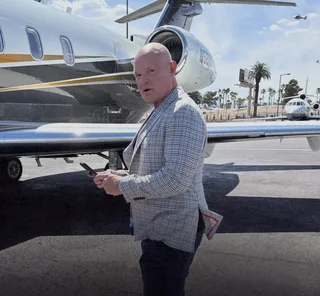 Shawn stands on a tarmac next to a private jet holding a phone.