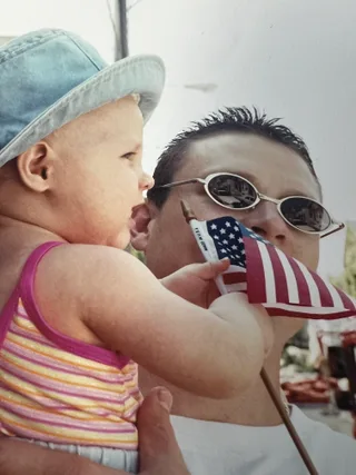 Shawn holds a baby outdoors while looking at a small American flag.