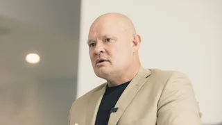 Shawn in a beige blazer and black shirt speaks while looking off-camera.