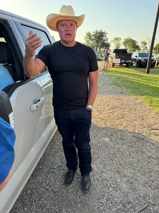 Shawn stands next to a truck wearing a cowboy hat and black t-shirt while gesturing with one hand.