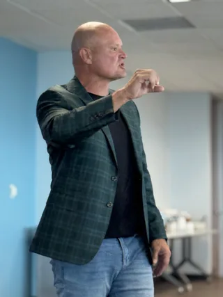 Shawn gestures with one hand while speaking during a presentation in a bright room.