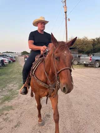 Shawn rides a brown horse outdoors near parked vehicles.