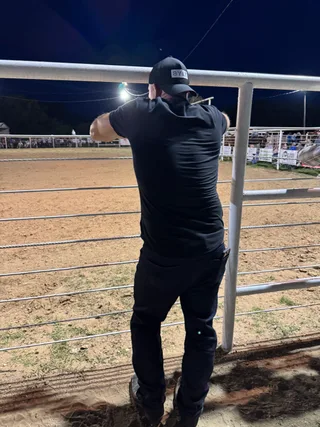 Shawn stands facing away from the camera, leaning against a white metal fence at a rodeo arena at night.