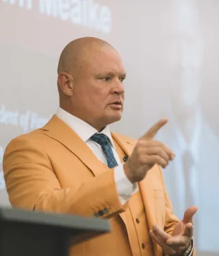 Shawn in an orange suit gestures while speaking at a podium.