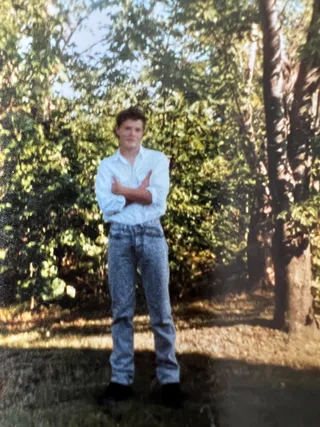 Shawn stands with arms crossed in front of trees.