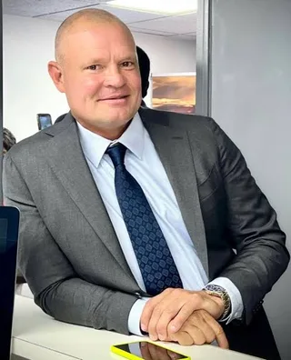 Shawn in a grey suit and blue tie leans on a white counter in an office setting.