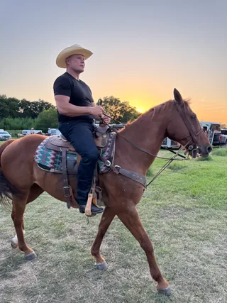 Shawn rides a horse outdoors in a field during sunset.