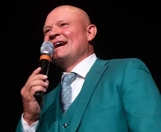 Shawn in a green suit and tie speaks into a microphone against a dark background.