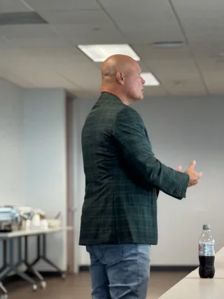 Shawn in a patterned blazer and jeans gestures while speaking in an office.