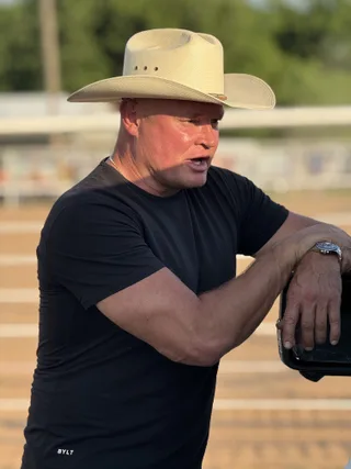 Shawn wears a cowboy hat and black t-shirt while leaning on a structure near an outdoor dirt track.