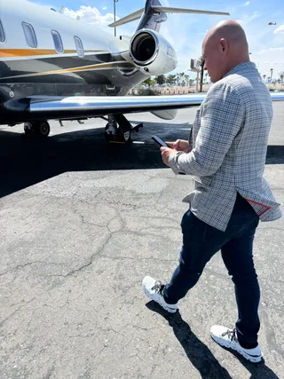 Shawn walks across an airport tarmac holding a smartphone near a private jet.