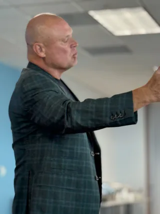 Shawn wears a plaid blazer and gestures while speaking in an office setting.