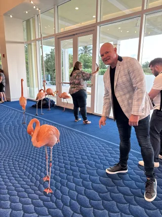 Shawn in a plaid blazer points toward a flamingo standing on a blue patterned carpet.