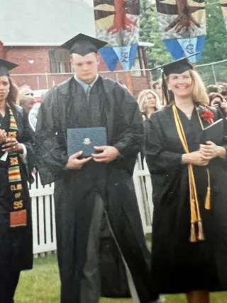 Shawn holds a diploma while wearing a cap and gown during a graduation ceremony outdoors.