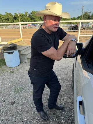 Shawn leans against a truck outdoors wearing a cowboy hat and black t-shirt.