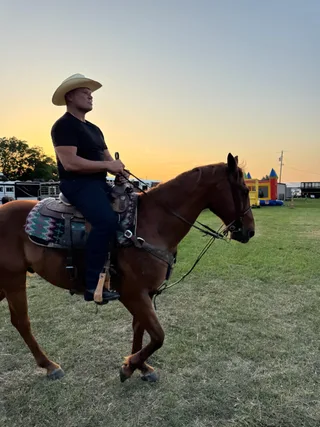 Shawn rides a brown horse outdoors during sunset wearing a cowboy hat.