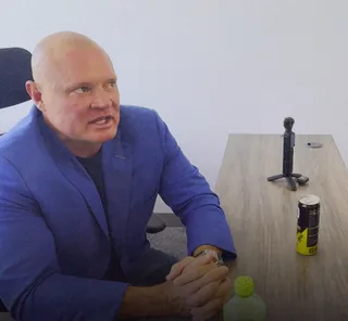 Shawn in a blue blazer sits at a wooden desk speaking while gesturing with clasped hands.