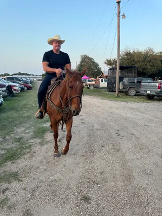 Shawn rides a brown horse along a dirt path outdoors near parked trucks.