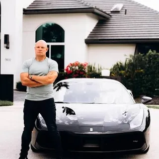 Shawn stands with arms crossed in front of a black sports car parked in a driveway.