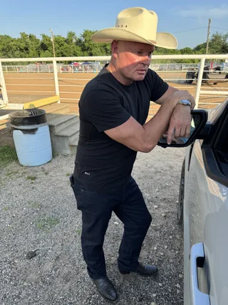 Shawn wearing a cowboy hat leans against a truck outdoors near a ranch.