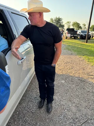 Shawn wearing a cowboy hat and black t-shirt stands next to a truck outdoors.