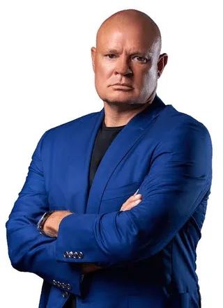 Shawn in a blue suit jacket stands with arms crossed against a white background.
