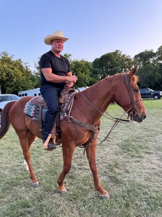 Shawn wears a cowboy hat while riding a brown horse across a grassy field.